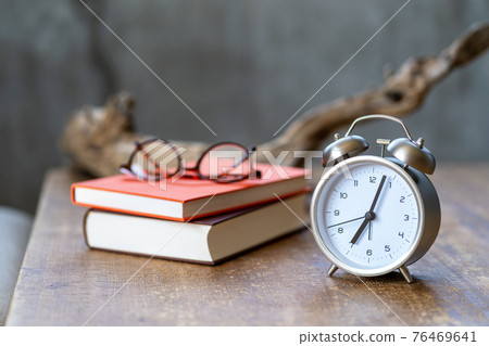An alarm clock, glasses, a book, and a dead tree lit by the rising sun placed on the side table 76469641