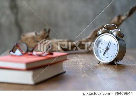 An alarm clock, glasses, a book, and a dead tree lit by the rising sun placed on the side table An alarm clock, glasses, a book, and a dead tree lit by the rising sun placed on the side table 76469644