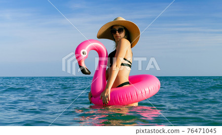 Resort wellness. Happy young sexy girl in bikini swimsuit, sunglasses and straw hat with pink inflatable flamingo in blue sea water on ocean beach background. On resort holiday vacation. 76471045