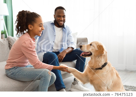 Smiling black girl playing with dog in the living room 76475123