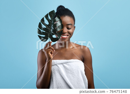 Pretty black woman with smooth skin wrapped in towel hiding behind tropical plant on blue studio background Pretty black woman with smooth skin wrapped in towel hiding behind tropical plant on blue studio background 76475148