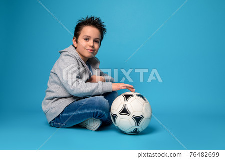 Handsome child sitting near a soccer ball on blue surface 76482699