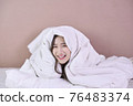 Cheerful young woman lying on bed playing with blanket and looking at camera. Asian girl feeling fresh after nap on bed. Laughing woman having fun while embracing pillow and blanket. 76483374