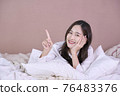 Beautiful woman wearing a white dress on bed showing and pointing something that is on her hand 76483376