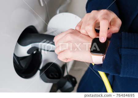 Woman uses her smart watch for opening an electric car and unplugging EV. Disconnecting the plug from charging the electric vehicle 76484695