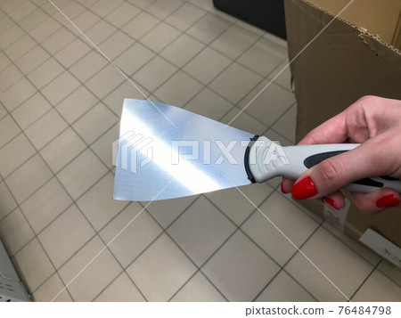 scraper for applying mastic, plaster. Painting works. a scraper in the hands of a woman with a bright red manicure. scraper with plastic handle. against the background of boxes in the store scraper for applying mastic, plaster. Painting works. a scraper in the hands of a woman with a bright red manicure. scraper with plastic handle. against the background of boxes in the store 76484798