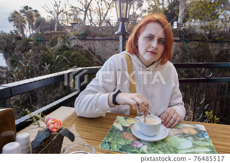 Terrace of restaurant by sea in Antalya, Turkey, young European red-haired woman relaxing at table with cup of coffee. 76485517