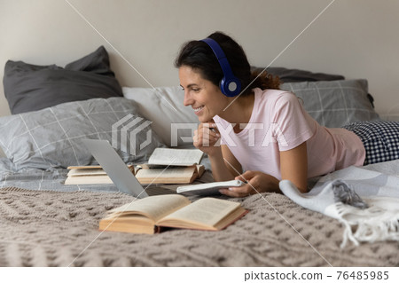 Millennial female wearing earphones lying on bed study using laptop 76485985