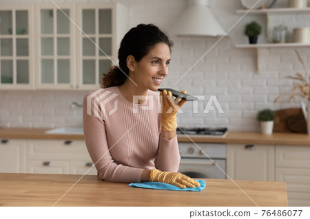Young latin housewife tidying up kitchen dictate message on phone 76486077
