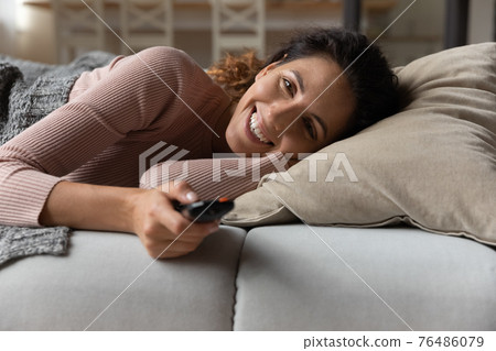 Happy latina female lie on cozy couch hold tv remote 76486079