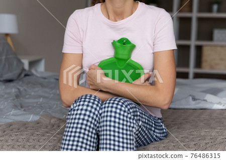 Woman sit on bed hold hot water bottle on belly Woman sit on bed hold hot water bottle on belly 76486315