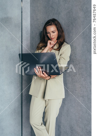 Woman stands at the wall and looks at her laptop screen Woman stands at the wall and looks at her laptop screen 76487649