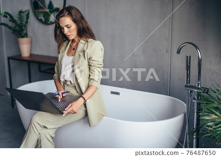 Woman sits on a bathtub in the bathroom and works with her laptop Woman sits on a bathtub in the bathroom and works with her laptop 76487650
