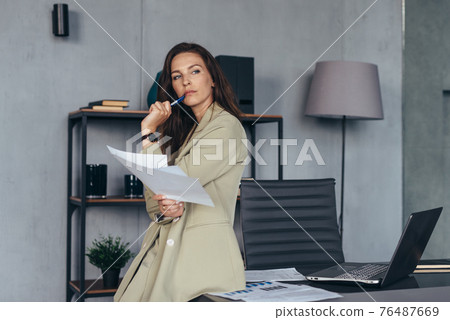 Woman stands thoughtfully with her papers leaning on her desk 76487669