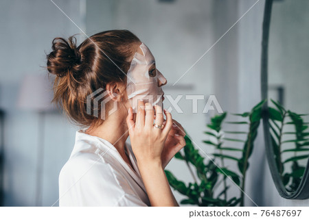 Woman grooming her face in the bathroom with a mask on her face in front of the mirror Woman grooming her face in the bathroom with a mask on her face in front of the mirror 76487697