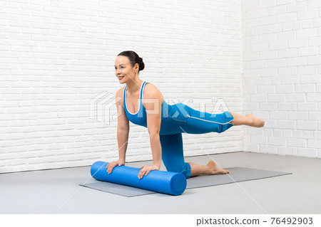 Pilates with props indoor. Fit smiling woman in blue sportswear practice bird dog drill with foam roller under her hands, one leg up, selective focus. 76492903