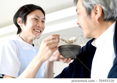 A young man in an apron helping a senior man with a meal A young man in an apron helping a senior man with a meal 76493526