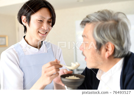 一個穿著圍裙的年輕人幫助一個老人吃飯 一個穿著圍裙的年輕人幫助一個老人吃飯 76493527
