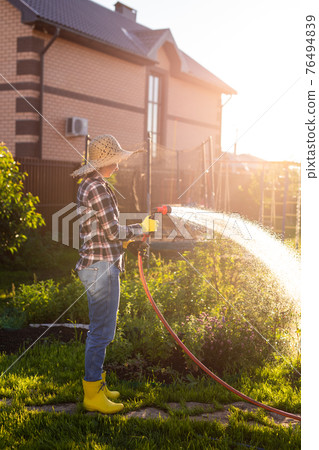 Caucasian woman gardener in work clothes watering the beds in her vegetable garden on sunny warm summer day. Concept of working in the garden and your farm 76494839