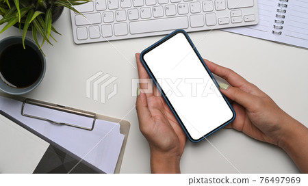 Above view of woman hands holding mobile phone with empty screen on whote office desk. Above view of woman hands holding mobile phone with empty screen on whote office desk. 76497969