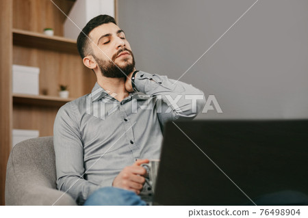 A handsome young man of oriental appearance works at home, sits in an armchair with a laptop and looks tired. 76498904