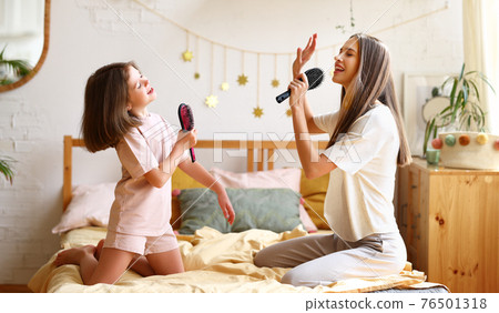 Enjoying weekend at home. Cute happy girl little daughter with hairbrush as mic singing with mom while sitting together on bed 76501318