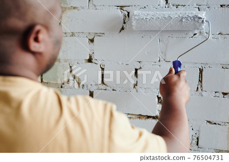 Hand of young African man in beige t-shirt holding paintroller while standing in front of brick wall Hand of young African man in beige t-shirt holding paintroller while standing in front of brick wall 76504731