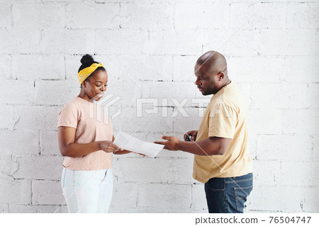 Young smiling African woman showing paper with photo of living-room interior to her husband Young smiling African woman showing paper with photo of living-room interior to her husband 76504747