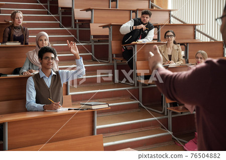 Young successful student in eyeglasses and casualwear raising hand and looking at professor 76504882
