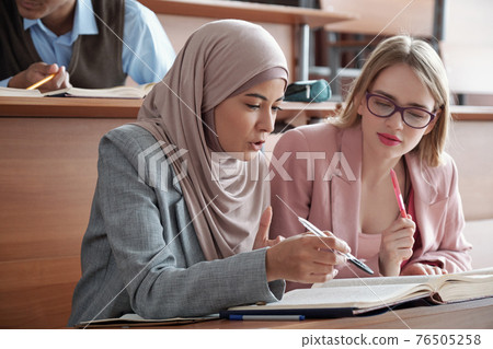 Two intercultural female students sitting by desk in lecture hall and discussing passage in book 76505258