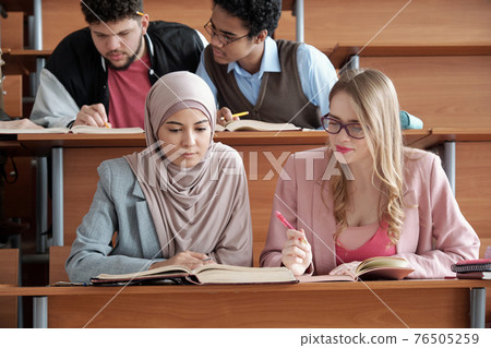 Two intercultural female students of university sitting by desk and looking through passage in book 76505259