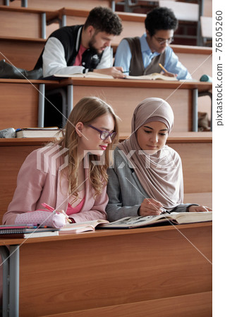 Two intercultural female students of university sitting on the first row by desk in lecture hall Two intercultural female students of university sitting on the first row by desk in lecture hall 76505260