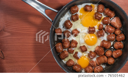 Banner with fried sausages with chicken eggs in process of frying in a frying pan at the red wood table with copy space for text. Banner with fried sausages with chicken eggs in process of frying in a frying pan at the red wood table with copy space for text. 76505462