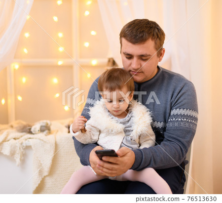 father and baby use smartphone, they play and watch device screen, home interior decorated with holiday lights and gifts father and baby use smartphone, they play and watch device screen, home interior decorated with holiday lights and gifts 76513630