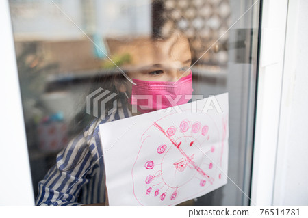 Sick small girl showing drawing through window at home, coronavirus concept. Sick small girl showing drawing through window at home, coronavirus concept. 76514781