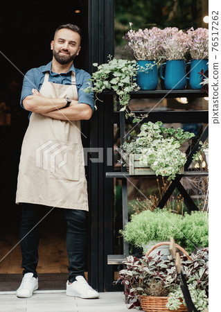 Cheerful millennial confident guy in apron with crossed arms in front door of flower shop 76517262