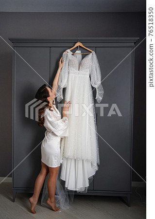 bride in a silk robe next to a white wedding dress hanging on the closet. bride in a silk robe next to a white wedding dress hanging on the closet. 76518350