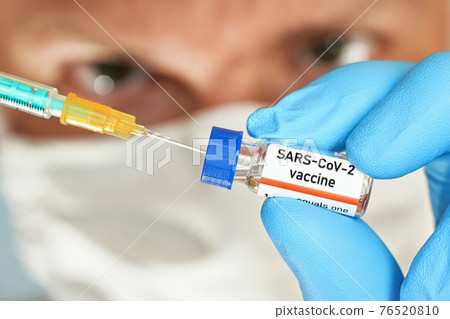 Man hand in blue glove hold small vial with coronavirus vaccine (label own design, not real product), to inject it with green orange syringe, blurred face with white virus mask in background 76520810