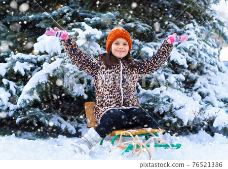 child girl sitting on a sled and playing with snow in the winter forest, bright snowy fir trees, beautiful nature 76521386