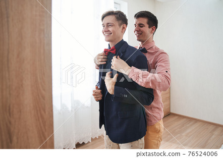 Gay man helping to put on a clothes to his boyfriend while standing with him Gay man helping to put on a clothes to his boyfriend while standing with him 76524006