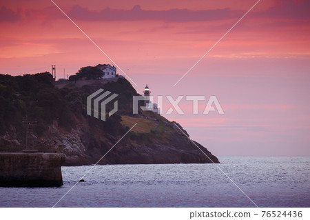 White lighthouse tower beaming on the rocky hill over the sea on the pink sunset White lighthouse tower beaming on the rocky hill over the sea on the pink sunset 76524436