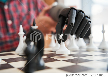 Engineer holding chess piece with black bionic hand while playing at the chess 76524850