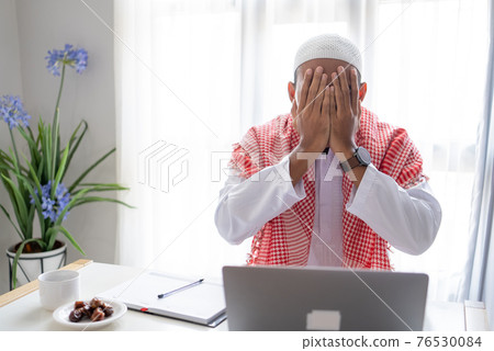 muslim man using laptop cover his face with hand during working from home 76530084