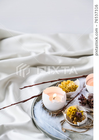 Trendy Easter decorations. Cracked organic eggs with dry grass flowers on plate. Candle light in White shell of the eggs. Bird feather. Copy space. Greeting easter card. Pastel colors. Trendy Easter decorations. Cracked organic eggs with dry grass flowers on plate. Candle light in White shell of the eggs. Bird feather. Copy space. Greeting easter card. Pastel colors. 76537316