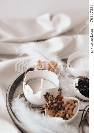Trendy Easter decorations. Cracked organic eggs with dry grass and dry flowers on plate. Candle in White shell of the eggs. Bird feather. Copy space. Greeting card. Pastel colors. Trendy Easter decorations. Cracked organic eggs with dry grass and dry flowers on plate. Candle in White shell of the eggs. Bird feather. Copy space. Greeting card. Pastel colors. 76537322