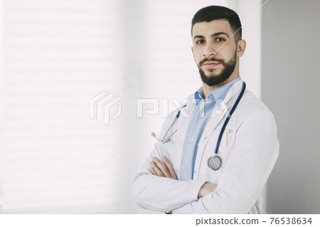 Serious young doctor in stethoscope and white medical coat stands in his office with arms crossed Serious young doctor in stethoscope and white medical coat stands in his office with arms crossed 76538634