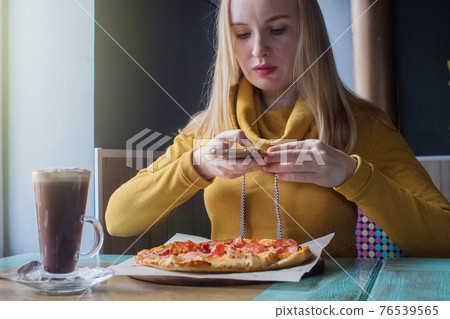 A blogger girl sits in a cafe and shoots a pizza on a smartphone. Work in social networks, video blogs and photo blogs. Concept for creating internet content and food photography. A blogger girl sits in a cafe and shoots a pizza on a smartphone. Work in social networks, video blogs and photo blogs. Concept for creating internet content and food photography. 76539565