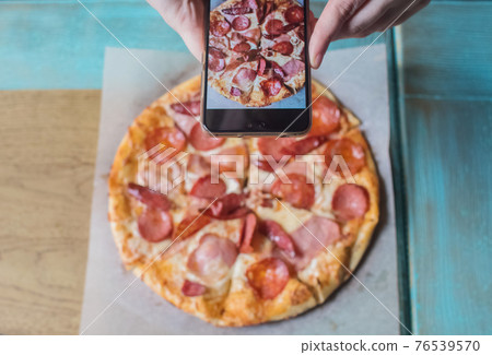A blogger girl sits in a cafe and shoots a pizza on a smartphone. Work in social networks, video blogs and photo blogs. Concept for creating internet content and food photography. A blogger girl sits in a cafe and shoots a pizza on a smartphone. Work in social networks, video blogs and photo blogs. Concept for creating internet content and food photography. 76539570
