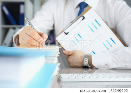 Businessman holds clipboard with commercial charts in his hands and makes notes in documents Businessman holds clipboard with commercial charts in his hands and makes notes in documents 76540495