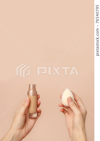 Applying foundation on makeup sponge. Woman's hands with neutral manicure holding bottle of concealer or toner foundation, cream and beauty blender, make up artist background 76540795